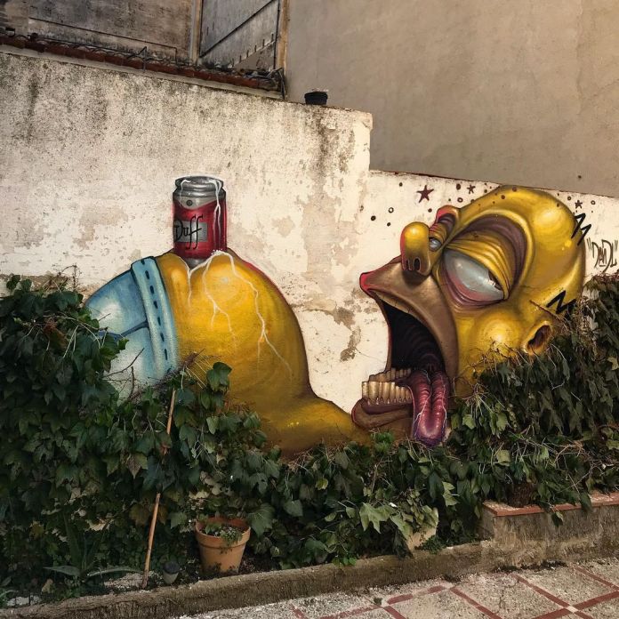 Post-graffiti artwork by DavidL featuring a surreal depiction of Homer Simpson with exaggerated features, holding a can of Duff beer, painted on a distressed wall with overgrown vegetation, located outside Barcelona, Spain.