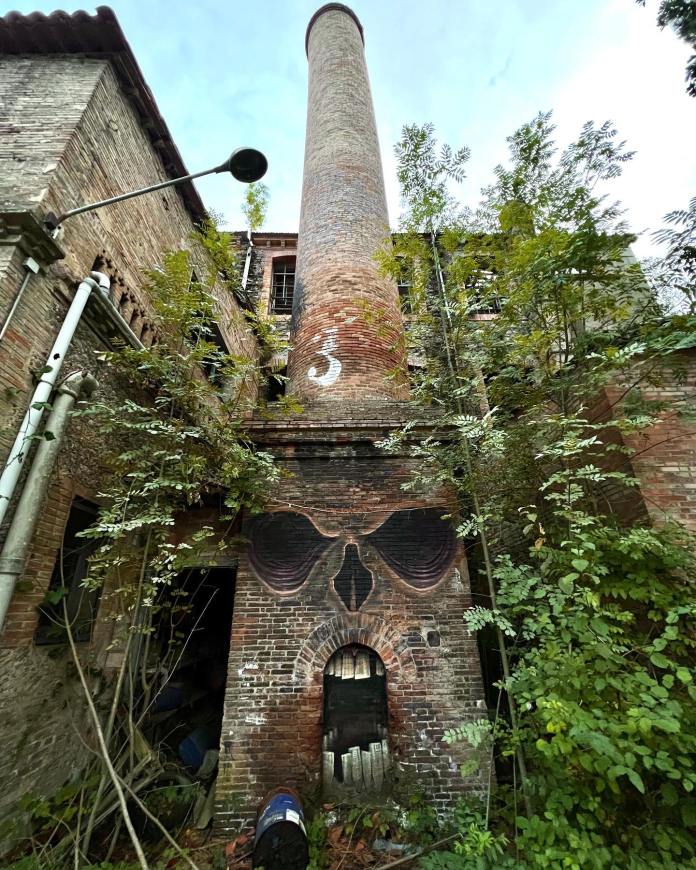 Post-graffiti artwork by DavidL featuring a skull face integrated into a brick chimney of an abandoned building, with overgrown vegetation surrounding the structure, located outside Barcelona, Spain.