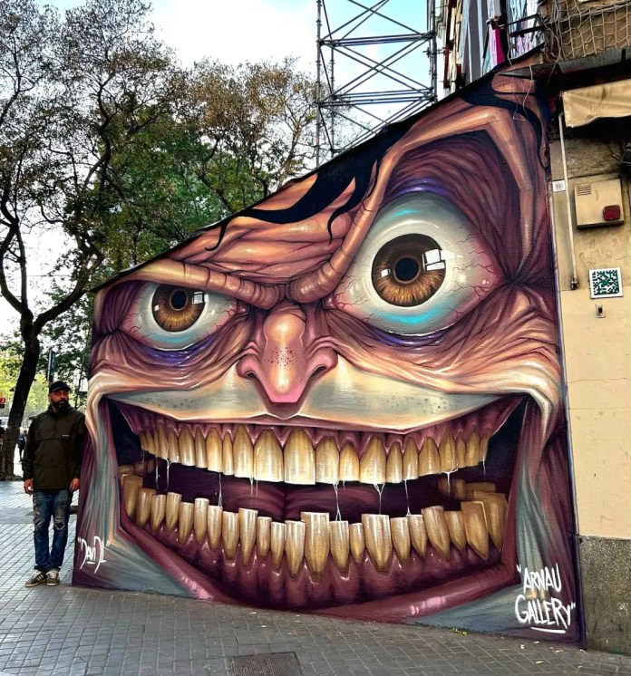 Post-graffiti artwork by DavidL featuring a large, exaggerated face with wide eyes and a menacing grin, painted on the exterior wall of a building in Barcelona, Spain, with the artist standing beside it.