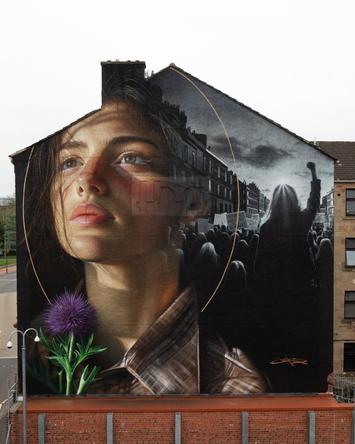 Photorealistic mural of a young woman with long hair and a calm but determined expression, facing left. Behind her is a grayscale city protest scene with a raised fist and placards. A bright purple thistle and golden arcs highlight the mural, which covers the entire side of a building in Glasgow, UK. Artwork by JEKS ONE for Yardworks.