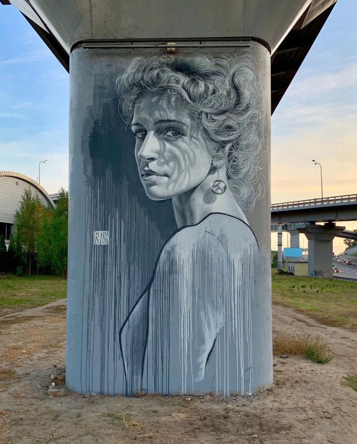 Monochromatic mural by Sasha Korban in Kyiv, Ukraine, painted on a concrete bridge pillar. The artwork features a realistic grayscale portrait of a woman with curly hair, wearing a dangling earring and gazing over her shoulder. Her expression is calm yet introspective, with subtle vertical paint drips blending into the texture of the pillar, creating a raw and emotional atmosphere.