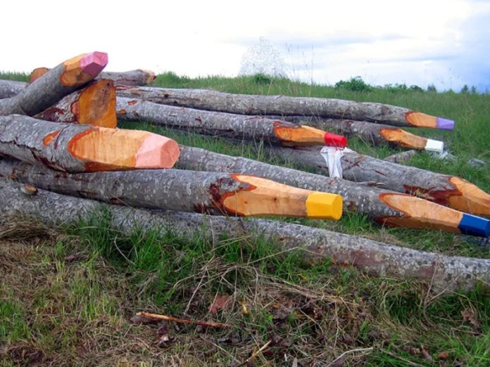 Tree logs carved and painted at the tips to look like oversized colorful pencils lying in a grassy field.