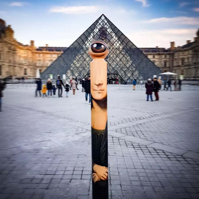 A painted bollard artwork by French artist Le CyKlop in front of the Louvre Pyramid in Paris, France. The cylindrical post is transformed into a playful reinterpretation of the Mona Lisa, featuring her folded hands, iconic face, and an oversized single cyclopean eye replacing her head. The background shows tourists and the glass pyramid entrance to the Louvre Museum, where the original Mona Lisa is housed.