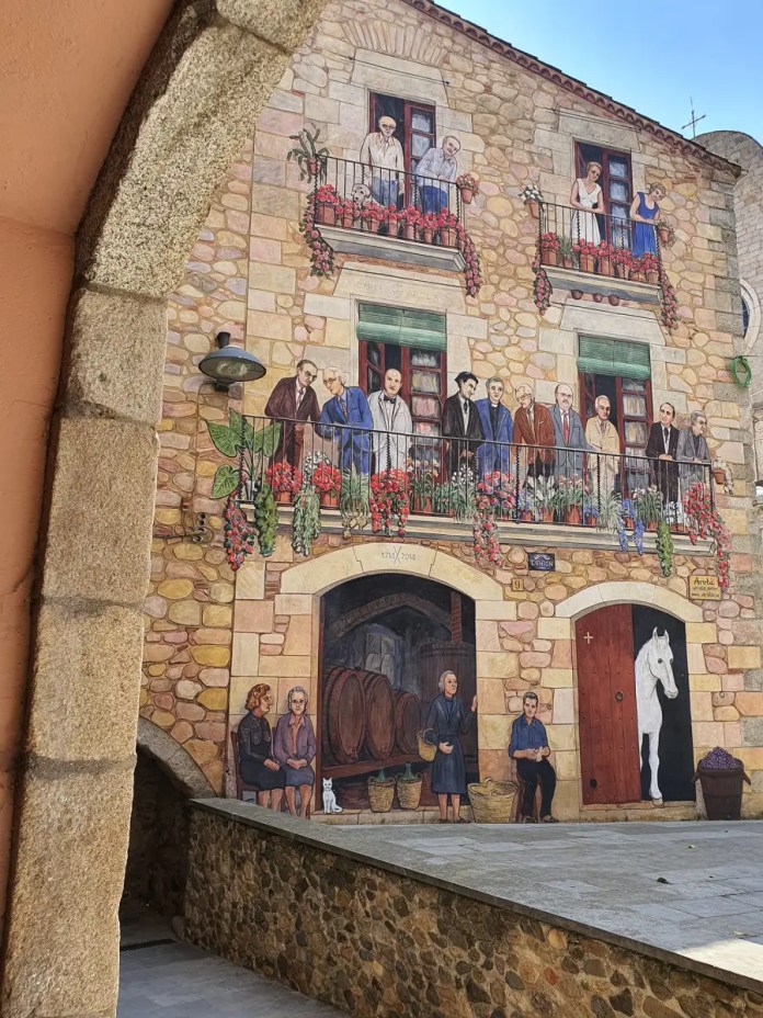 A stunning trompe-l'œil mural by Carles Arola in Calonge, Spain, covering the façade of a stone building. The painting features lifelike depictions of people on balconies surrounded by vibrant red flowers, an open stable door with a white horse, and ground-level scenes of elderly women, barrels of wine, and local market activity. The illusion blends seamlessly with the building’s architecture, showcasing elements of the town's cultural heritage.