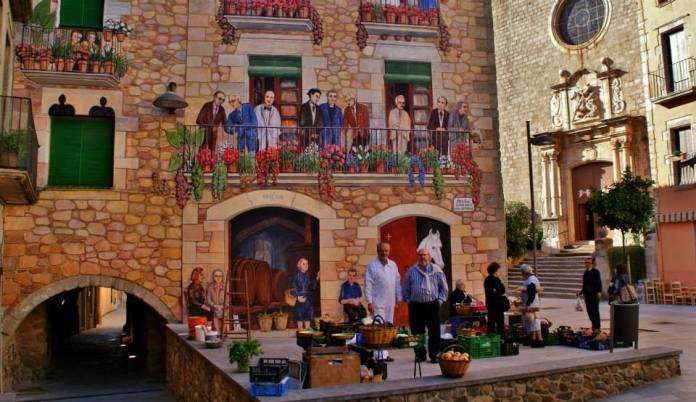 A beautifully detailed trompe-l'œil mural by Carles Arola on a stone façade in Calonge, Spain. The mural depicts lifelike scenes of people on flower-adorned balconies, a wine cellar with barrels, and a white horse emerging from a stable door. In front of the mural, a local woman sells fresh produce, adding a layer of real-life activity to the painted illusion. The juxtaposition of art and reality showcases the community’s vibrant cultural heritage.