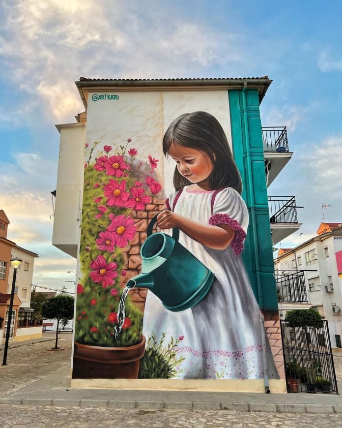 Mural of a young girl watering a pot of pink flowers on the side of a building in Ronda, Spain, painted by Kato Art. The girl wears a white dress and holds a blue watering can.