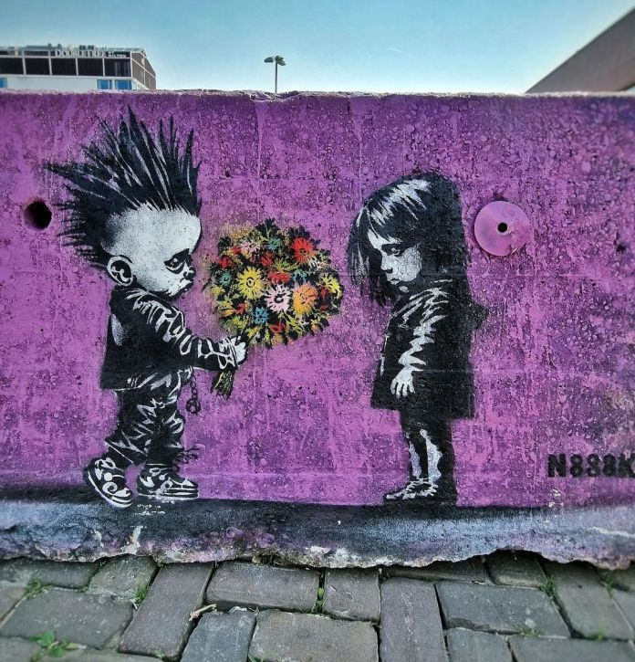 Street art on a purple wall showing a punk boy giving flowers to a sad girl, painted by N888K in Amsterdam. The scene uses black-and-white stencils with bright flowers.