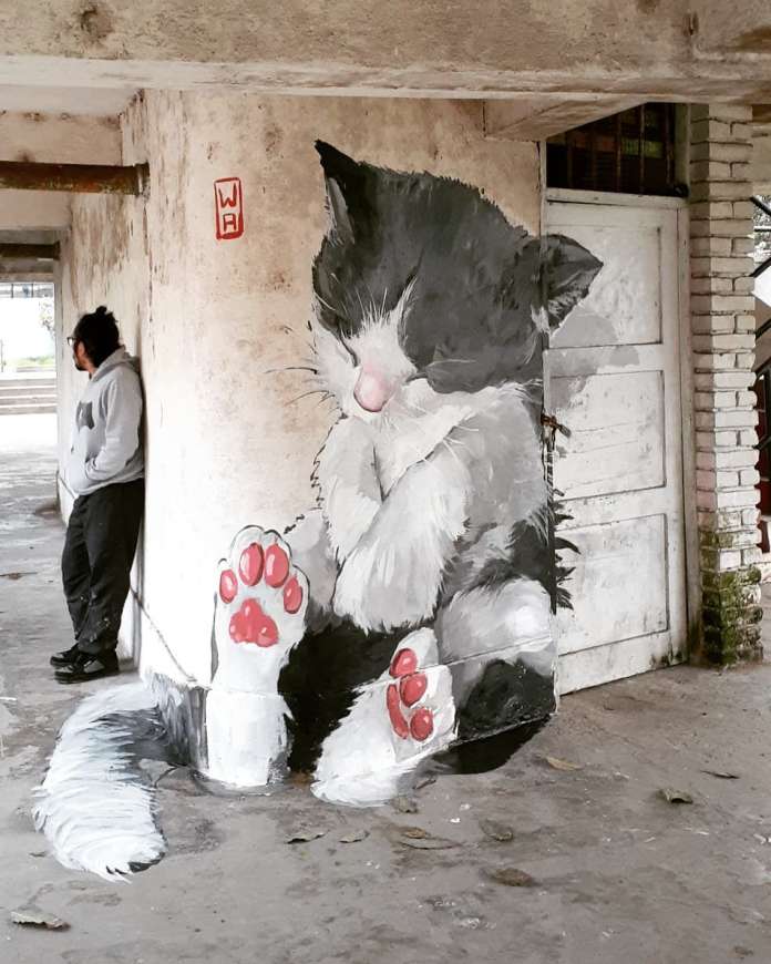 Mural of a black and white kitten curled up around a pillar in Lima, Peru. The kitten has pink paws and closed eyes, blending into the architectural corner.