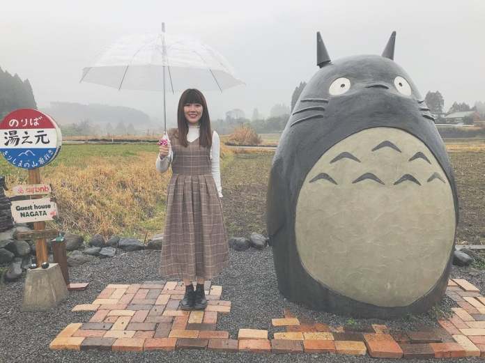 A life-size Totoro sculpture from Studio Ghibli's My Neighbor Totoro stands next to a smiling visitor holding a clear umbrella. The Totoro is gray with a round belly, surrounded by a neat brick and gravel path. Next to it is a red and blue bus stop sign, labeled "Guest House Nagata," located in Takaharu, Japan. The background shows a foggy rural landscape with fields and trees.