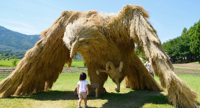 A large straw sculpture resembling a mythical bird with wide-spread wings at the Wara Art Festival in Niigata, Japan. A young child stands in front of the sculpture, highlighting its massive scale. The sculpture is set against a bright, sunny landscape with lush greenery and distant hills.