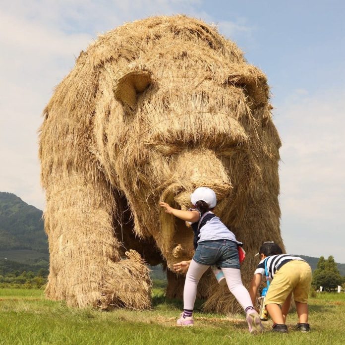 A giant straw sculpture of a bear at the Wara Art Festival in Niigata, Japan. Two children are interacting with the sculpture, one reaching towards its nose and the other crouching nearby, showing its playful and interactive nature. The bear's detailed features and the green field with a blue sky in the background create a lively outdoor scene.