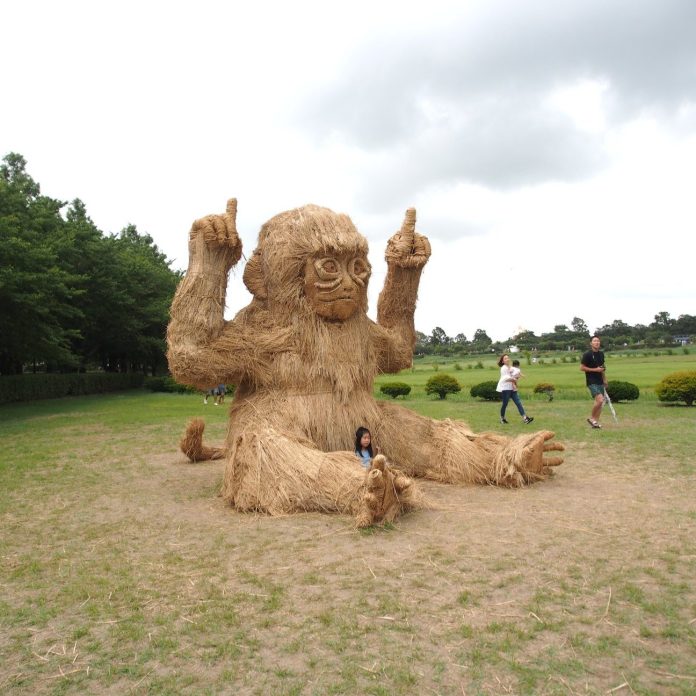 A giant straw sculpture of a sitting monkey at the Wara Art Festival in Niigata, Japan. The monkey has a playful expression with raised hands, and a child sits comfortably in its lap, emphasizing the interactive and family-friendly atmosphere of the festival. The open grassy area and cloudy sky create a relaxed outdoor setting.