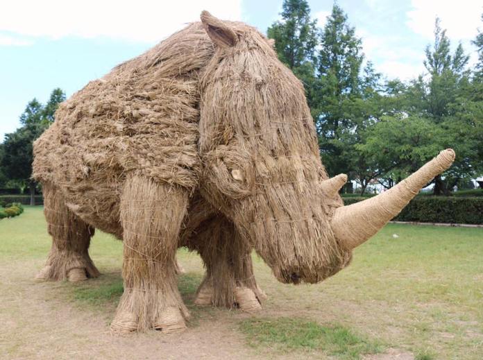 A giant straw sculpture of a rhinoceros at the Wara Art Festival in Niigata, Japan. The sculpture features detailed textures and a large horn, standing on a grassy field surrounded by trees and a clear sky. The artwork highlights the creativity and craftsmanship involved in the festival.