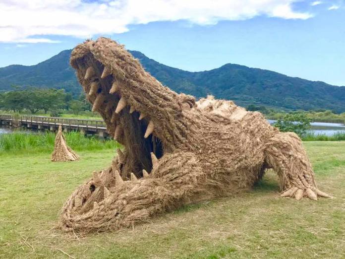 A giant straw sculpture of a crocodile with its mouth wide open, showcasing detailed teeth, at the Wara Art Festival in Niigata, Japan. Set on a grassy field with mountains and a river in the background, the sculpture blends creativity with the natural surroundings, creating a striking scene.
