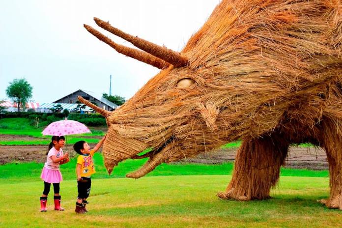 wo children interact with a giant straw sculpture of a triceratops at the Wara Art Festival in Niigata, Japan. One child, holding a pink umbrella, stands nearby while the other touches the sculpture's face, highlighting its interactive nature. The vibrant green field and a farmhouse in the background enhance the festive setting.
