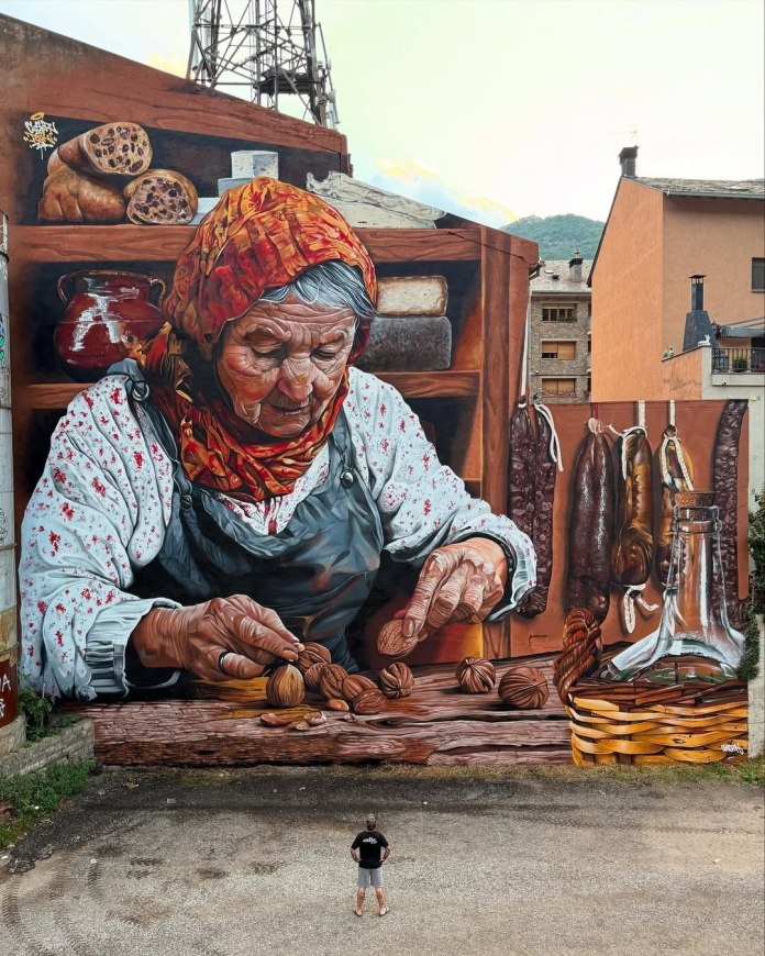 Large hyperrealistic mural of an elderly woman with a red scarf and floral blouse cracking walnuts, surrounded by hanging sausages, loaves of bread, cheese, and traditional pantry items. Painted on a tall exterior wall in Sort, Spain.