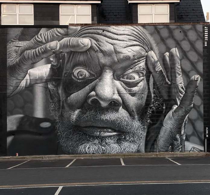 Black-and-white mural of an older man staring with widened eyes and pulling his eyelids apart with his fingers. Painted in photorealistic style across a wall with windows at the top.