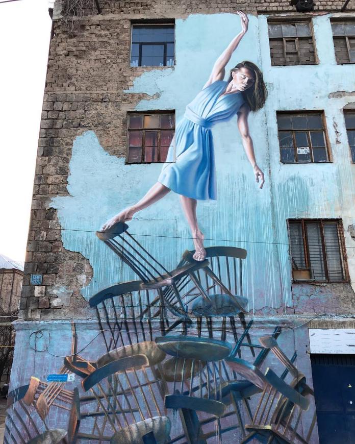 Mural of a woman in a blue dress balancing on unstable wooden chairs painted against a weathered brick wall, with her arms outstretched as if walking a tightrope.