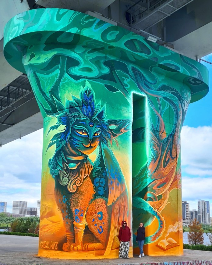 Vibrant mural on a bridge column depicting a mystical feline with glowing eyes, a third eye, and vivid orange, green, and blue fur. Stylized roots and branches wrap around the cylindrical surface.
