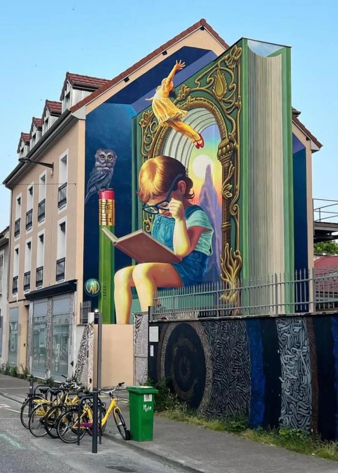 Mural of a young child reading in front of an open book painted as an architectural arch. A girl leaps from the pages, and an owl sits on a pencil beside the scene. The book’s spine adds dimensional realism.