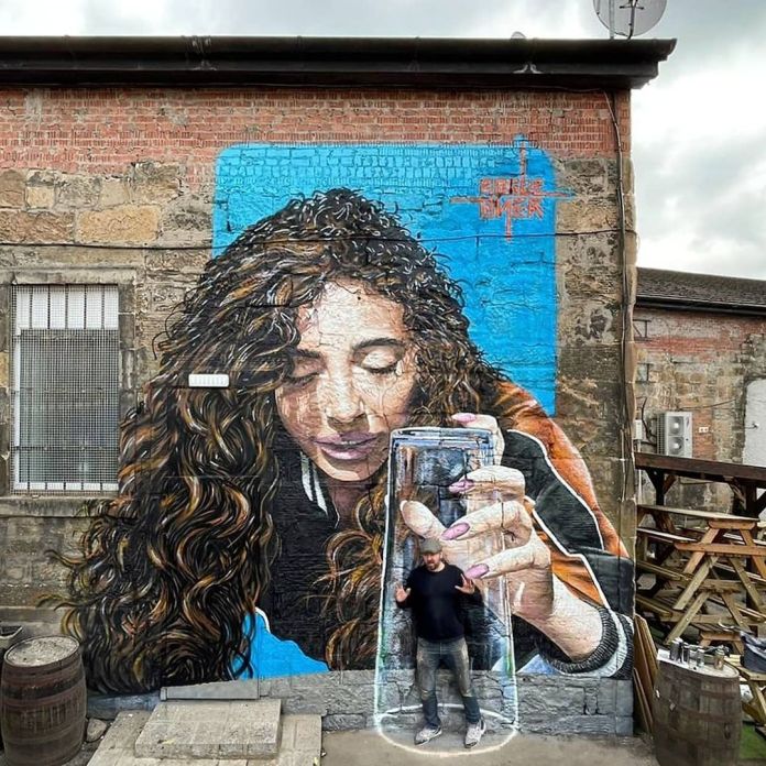 Mural of a woman with long curly hair gently lowering a glass over a real man standing in front of the wall. The man appears trapped inside the painted glass. Located on a brick building in Glasgow, UK, with the illusion completed by the viewer's position.