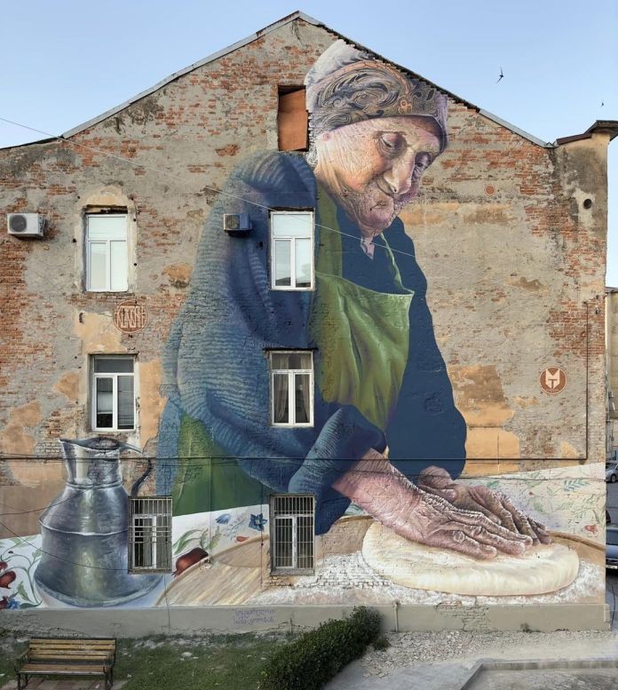 Large mural of an elderly woman baking bread, her hands working dough across a long wall, set against a brick façade with embedded windows in Kutaisi, Georgia.
