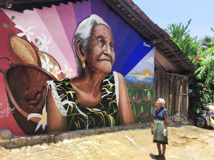Bright mural of an elderly woman against a radiant purple-to-blue background, with cocoa pods and floral details representing Mexican culinary tradition.
