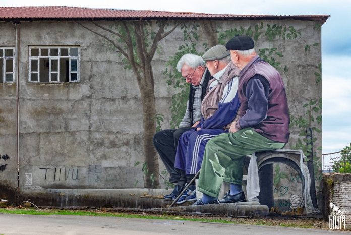 Mural of three elderly men sitting on a low concrete ledge, casually chatting in front of a cracked wall with tree shadows painted behind them.