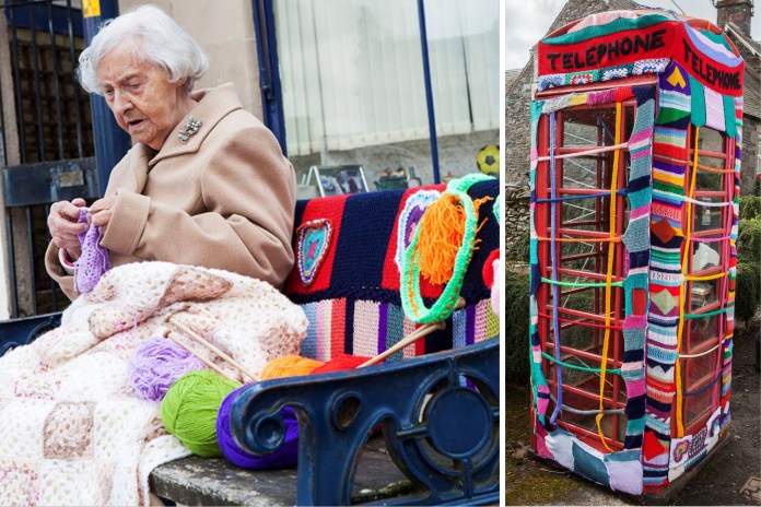 Elderly woman knitting on a bench covered in colorful yarn, with skeins and patchwork surrounding her, next to a British phone booth wrapped in crochet.