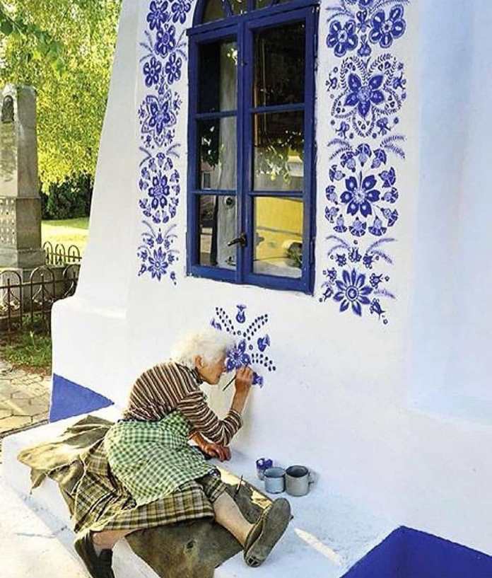 An elderly woman, Anežka Kašpárková, sits on a low bench, meticulously painting intricate blue floral designs around a blue-framed window on a white wall. Dressed in a plaid skirt, striped shirt, and green checkered apron, she brings traditional Moravian patterns to life, enhancing the charm of her Czech village. The scene is bathed in soft sunlight, highlighting her dedication to transforming her community into an open-air gallery.