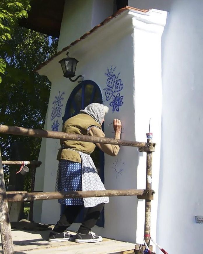 Anežka Kašpárková, a 90-year-old Czech artist, stands on a wooden scaffold, hand-painting intricate blue floral patterns onto the white walls of a village building. Dressed in a layered outfit with a blue-patterned apron, headscarf, and sneakers, she leans carefully to perfect her traditional Moravian-style artwork near a blue-framed arched window. A rustic lamp hangs above her as she transforms the plain walls into vibrant works of art, highlighting her dedication to preserving and celebrating local culture.