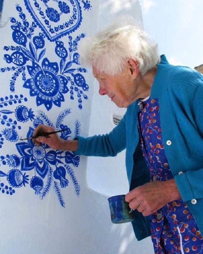 Anežka Kašpárková, a 90-year-old Czech artist, is seen up close as she meticulously paints vibrant blue floral patterns on a white wall, holding a small brush in one hand and a paint cup in the other. Wearing a patterned dress and a bright blue cardigan, she adds intricate details to her traditional Moravian-inspired designs. Her focused expression highlights her passion and precision as she transforms the plain wall into a stunning piece of folk art, celebrating her village's cultural heritage.