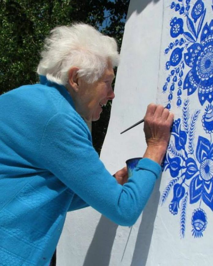Anežka Kašpárková, a 90-year-old Czech artist, smiles gently as she adds intricate blue floral patterns to a white wall in her village. Wearing a bright blue cardigan, she holds a small brush in one hand and a paint cup in the other, carefully bringing the traditional Moravian designs to life. Her work, a beautiful celebration of her cultural heritage, is framed by the surrounding greenery, emphasizing her dedication to transforming her community into an open-air gallery.