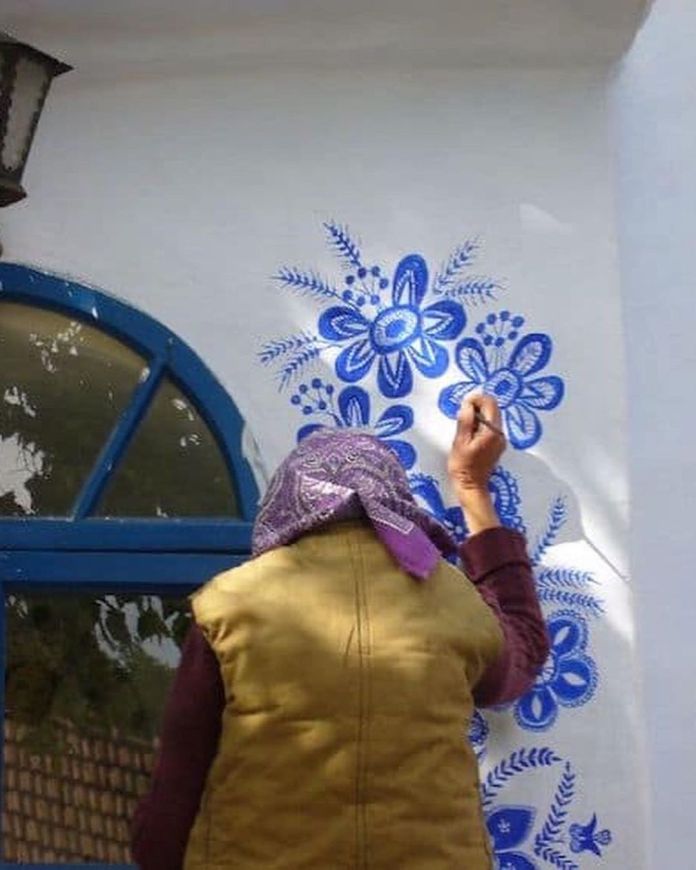 Anežka Kašpárková, a 90-year-old Czech artist, is seen from behind as she carefully paints vibrant blue floral patterns on a white wall near an arched blue-framed window. Wearing a purple-patterned headscarf, a beige vest, and a burgundy shirt, she skillfully adds intricate details to the traditional Moravian-inspired designs. Her work highlights her dedication to transforming the buildings in her village into stunning displays of cultural artistry.