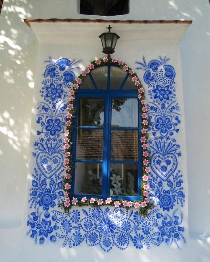 A beautifully painted arched window framed in vibrant blue with intricate floral patterns in traditional Moravian style, hand-painted by Anežka Kašpárková, a 90-year-old Czech artist. The white wall is adorned with delicate blue flowers, hearts, and ornate details, while the window itself is surrounded by a garland of pink and white artificial flowers. Above the window, a rustic black lamp with a cross adds a charming touch to this unique artwork, reflecting both cultural heritage and artistic passion.