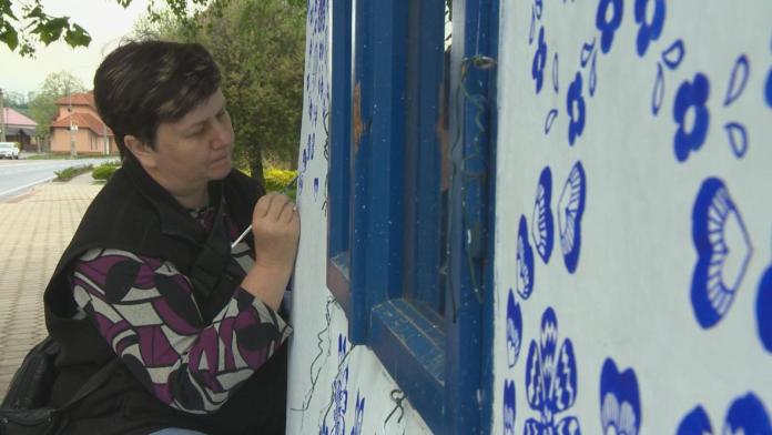 Marie Jagošová, a Czech artist, leans closely to a white wall as she paints intricate blue floral and heart patterns near a blue-framed window of a chapel in Louka. Dressed in a patterned shirt and black vest, she carefully adds traditional Moravian designs to the building. The background shows a quiet village street lined with greenery and houses, highlighting the serene setting of her artistic contribution to the community.