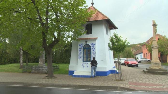 A small white chapel in Louka, Czech Republic, adorned with vibrant blue floral patterns surrounding its arched window, stands as a unique artistic landmark. The chapel features a terracotta-tiled roof and a blue-painted base, adding to its charm. A tree and a historic cross are visible nearby, while Marie Jagošová works on the wall, continuing the tradition of hand-painting intricate Moravian designs. The surrounding area includes a quiet street lined with greenery and parked cars, reflecting the serene village setting.