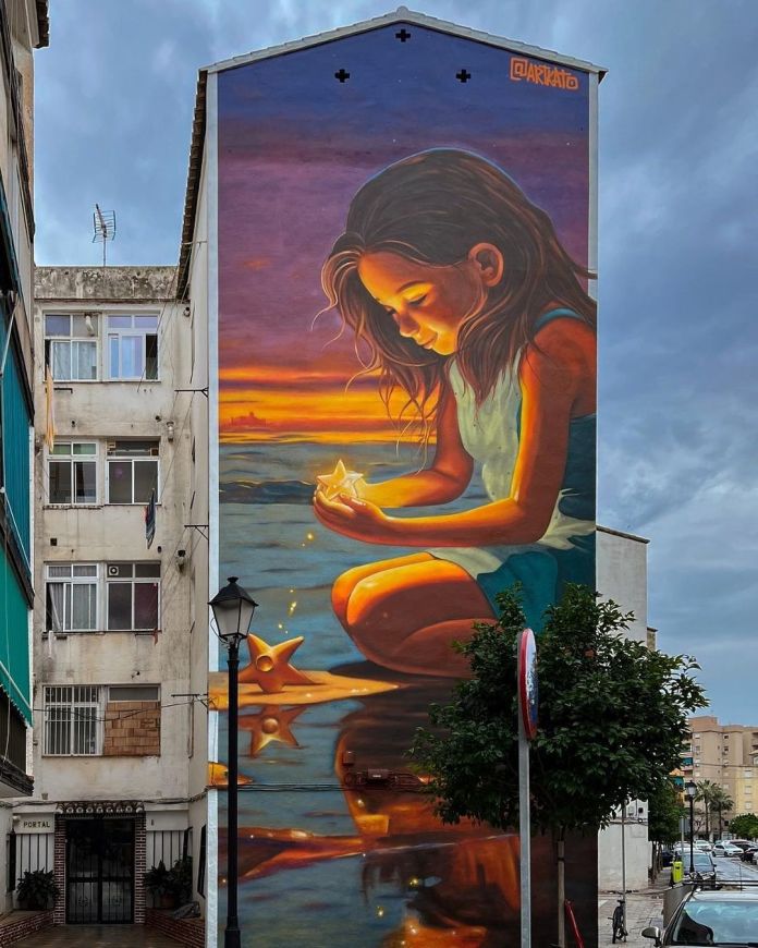 Large mural of a girl kneeling on a beach at sunset, holding a glowing origami star. Warm golden tones reflect off the water as she gazes at the star in quiet focus.