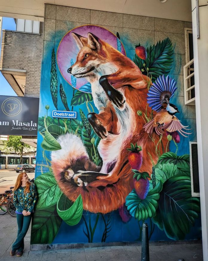 Vibrant mural titled 'Stadsnatuur' by artist Nina Valkhoff in Rotterdam, Netherlands. The artwork features a red fox surrounded by lush green plants, strawberries, and birds, capturing a blend of urban nature. The fox appears to be in motion, with its fur flowing and eyes alert. A small bird with colorful wings adds an extra touch of whimsy to the scene. The artist stands beside the mural, providing a sense of scale. This piece brings an enchanting glimpse of wildlife to the urban streetscape.