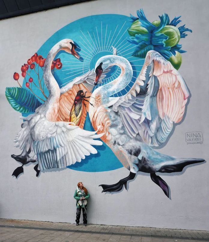 Mural titled 'The Irish Mythological Story of Étaín' by artist Nina Valkhoff for the DRAWDA festival in Drogheda, Ireland. The artwork features two majestic swans entwined, symbolizing Étaín and her lover in the myth, set against a circular blue background. A vibrant scarlet fly—Étaín's cursed form—appears between them, glowing with intricate detail. Surrounding elements include vivid green leaves and red berries, adding a natural touch to the mythological scene. The artist stands below, giving a sense of scale to this visually stunning tribute to Irish folklore.