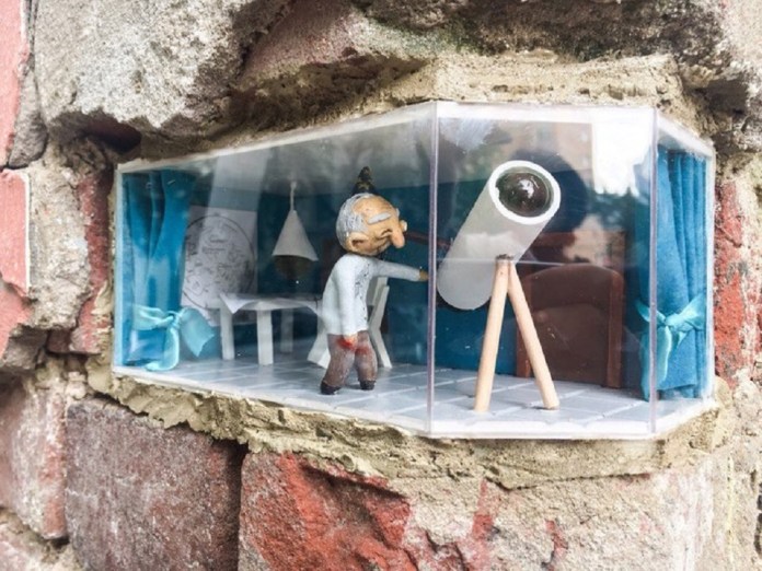 Close-up of a miniature street art installation by Ivan Sery in Nizhny Novgorod, Russia, featuring a tiny room embedded into a brick wall. The scene showcases a figure of an elderly man with a white coat and a magnifying glass, standing beside a telescope. The room is adorned with blue curtains, a small table, and a lamp, creating a whimsical and detailed diorama that blends seamlessly with the urban environment.