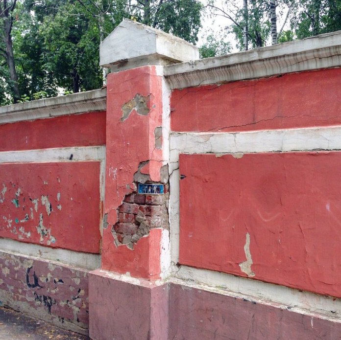 A street art installation by Ivan Sery embedded into a cracked red and white plaster wall in Nizhny Novgorod, Russia. The miniature diorama is nestled in an exposed brick section of the wall, surrounded by weathered and peeling paint. The small art piece adds an element of surprise and creativity to the deteriorating urban structure, blending seamlessly into the environment.