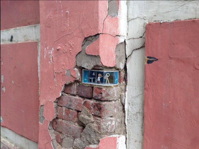 Close-up of Ivan Sery's miniature street art installation in Nizhny Novgorod, Russia, embedded in a weathered red and white plaster wall with exposed bricks. The tiny diorama depicts a whimsical scene of an elderly man with a telescope, standing in a detailed room complete with blue curtains and furniture. The artwork blends seamlessly into the cracked wall, creating a striking contrast between the urban decay and the playful creativity of the installation.