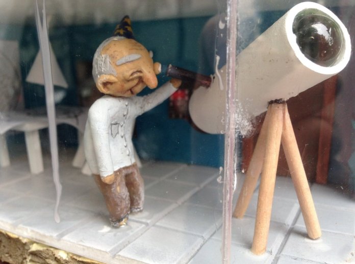 Extreme close-up of Ivan Sery's miniature diorama embedded in a brick wall in Nizhny Novgorod, Russia. The artwork features a detailed figurine of an elderly man wearing a white coat, peering through a small telescope mounted on a tripod. The scene includes intricate details such as a tiled floor, blue walls, and miniature furniture, emphasizing the artist's meticulous craftsmanship and creative integration into the urban environment.