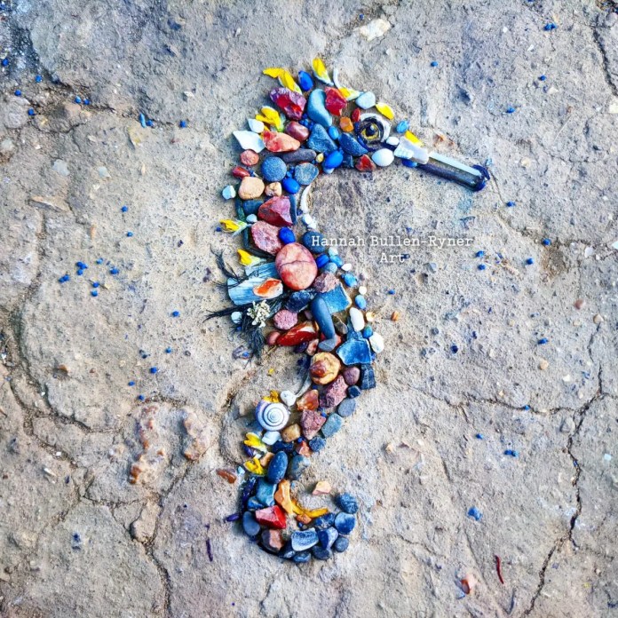 A seahorse created by Hannah Bullen-Ryner using a collection of tiny natural materials arranged intricately on the ground. The seahorse’s body is crafted from a variety of small stones, pebbles, and colorful elements, including red, blue, and yellow petals, shells, and bits of organic debris. The mix of vibrant and earthy tones captures the delicate form of a seahorse with careful attention to detail. Each piece is purposefully selected, demonstrating the artist's meticulous search for materials and dedication to reusing and preserving her collection. This ephemeral artwork reveals the artist’s unique process, bringing a whimsical, miniature ocean creature to life in a natural setting.