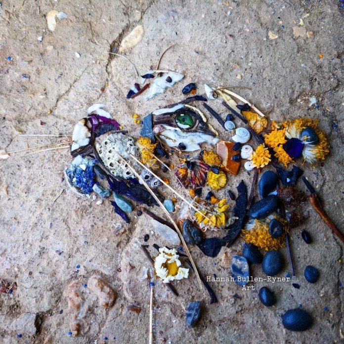 An ephemeral artwork by Hannah Bullen-Ryner depicting a leopard, crafted from an array of natural materials arranged on the ground. The leopard’s face is intricately detailed, with a striking green eye made from a small leaf or stone, and surrounding elements of yellow and orange flowers to represent the fur’s vibrant spots. Twigs, seeds, and stones form the facial structure, while delicate petals and tiny shells add texture and dimension. The organic materials come together to capture the leopard’s fierce yet gentle expression, showcasing the artist’s skill in creating lifelike representations from nature’s ephemeral elements.