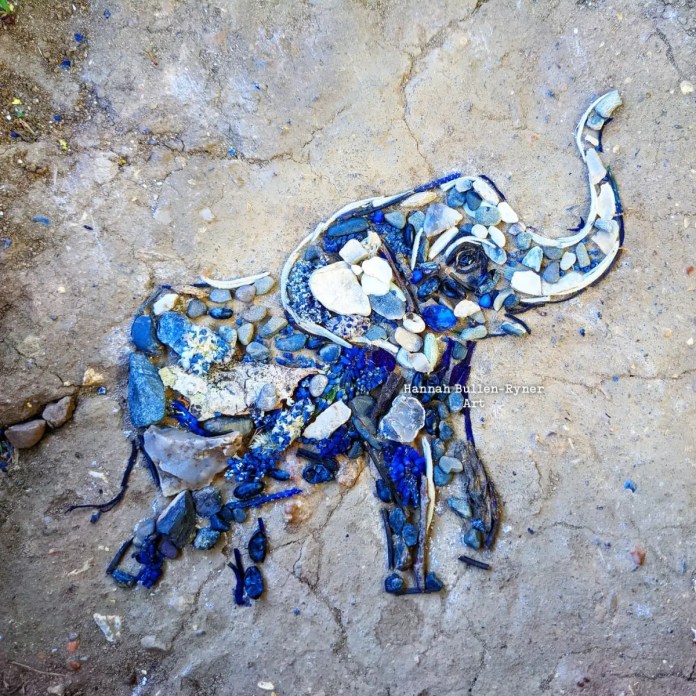 An ephemeral artwork by Hannah Bullen-Ryner depicting a baby elephant, crafted from an assortment of natural materials laid on the ground. The elephant’s form is created using small stones, twigs, and pebbles in shades of blue, gray, and white, carefully arranged to capture the gentle contours of its body and upturned trunk. The artist has included a dark stone to highlight the elephant’s eye, adding a lifelike touch to the figure. The use of cool tones and earthy textures reflects the strength and grace of this young elephant, brought to life through a temporary yet charming arrangement of organic elements.