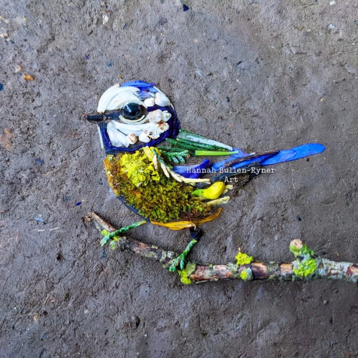 An ephemeral artwork by Hannah Bullen-Ryner depicting a tiny Bluetit, crafted from natural materials arranged on the ground. The bird’s body features a vibrant, mossy green and yellow "tummy," representing the distinctive colors of a Bluetit, while its head is formed from white and blue petals. A small black stone creates a sparkling eye, capturing the bird’s lively expression. Perched on a twig covered in lichen, the Bluetit is delicately assembled, with each feather hinted at through small leaves and petals. This piece is a symbol of love and hope, dedicated to those affected by war, showcasing nature’s beauty and resilience.