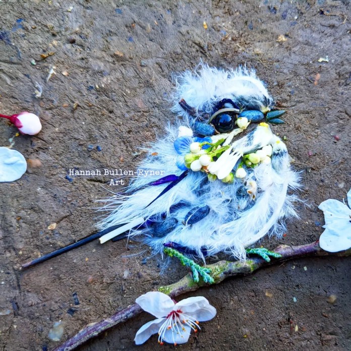An ephemeral artwork by Hannah Bullen-Ryner depicting a fluffy, small bird, crafted spontaneously from natural materials on the ground. The bird's soft, feathery body is created with delicate white petals, fuzzy seed pods, and small stones, giving it a downy appearance. A dark, shiny stone forms the eye, surrounded by tiny leaves and blooms, capturing the gentle, inquisitive look of the bird. Perched on a lichen-covered twig with scattered blossoms nearby, this piece radiates warmth and charm, showing the artist’s intuitive approach in a short, free-flowing session. The result is a heartwarming, whimsical bird that brings a smile to the viewer.