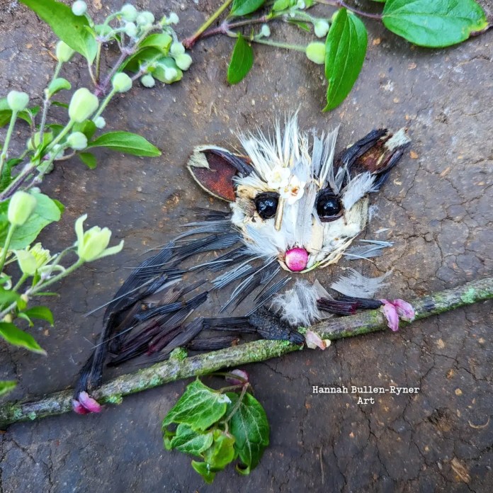 An ephemeral artwork by Hannah Bullen-Ryner depicting a tiny, baby opossum crafted from natural materials arranged on the ground. The opossum’s face is charmingly detailed with shiny dark stones for eyes, a small pink petal for the nose, and light-colored leaves and feathers for fur. The creature clings to a twig, with its tiny “paws” made from pink petals, adding a playful, endearing touch. Surrounding greenery frames the piece, enhancing the woodland ambiance. This creation captures the innocence and sweetness of a baby opossum, crafted in a temporary, nature-inspired form that seems ready to be carried away in a pocket.